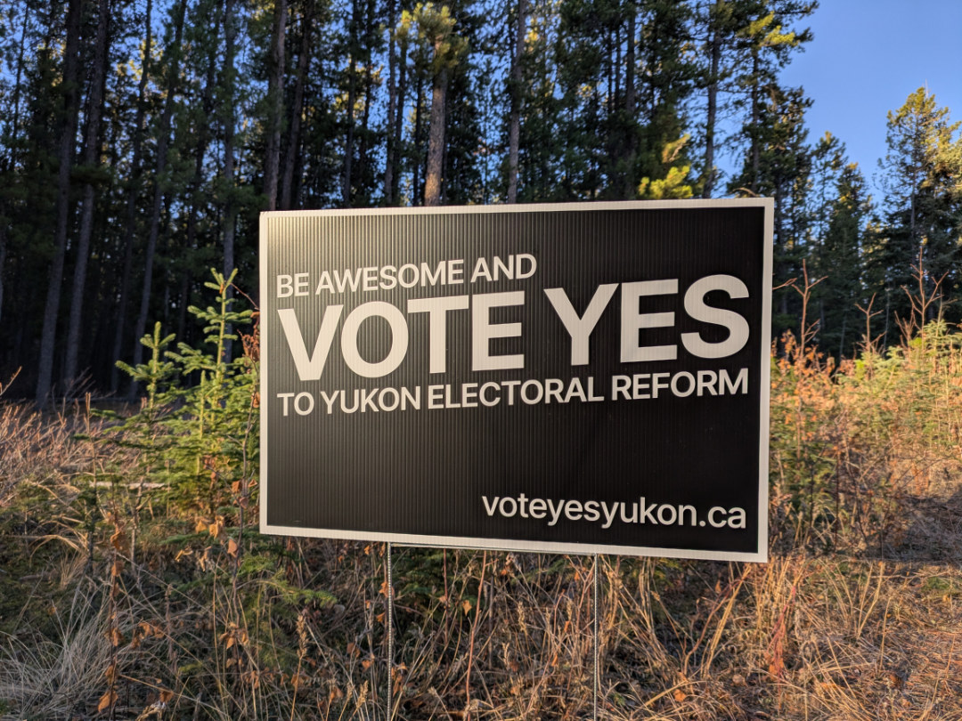 A “Vote YES Yukon” lawn sign in front of some pine trees near a Whitehorse trail.