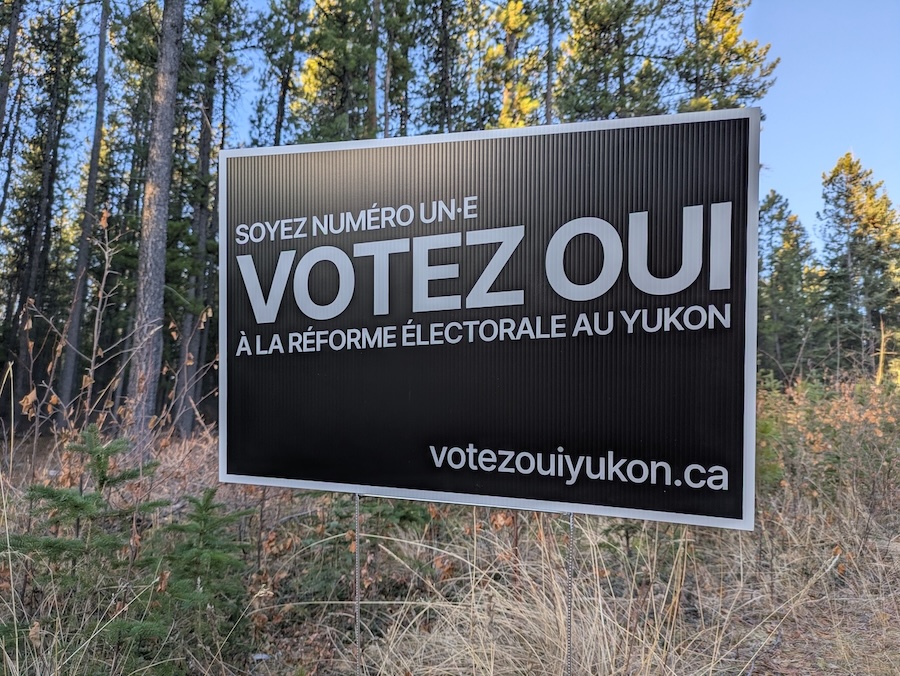 A “Votez OUI Yukon” sign in front of some pine trees, with the sun shining on it.