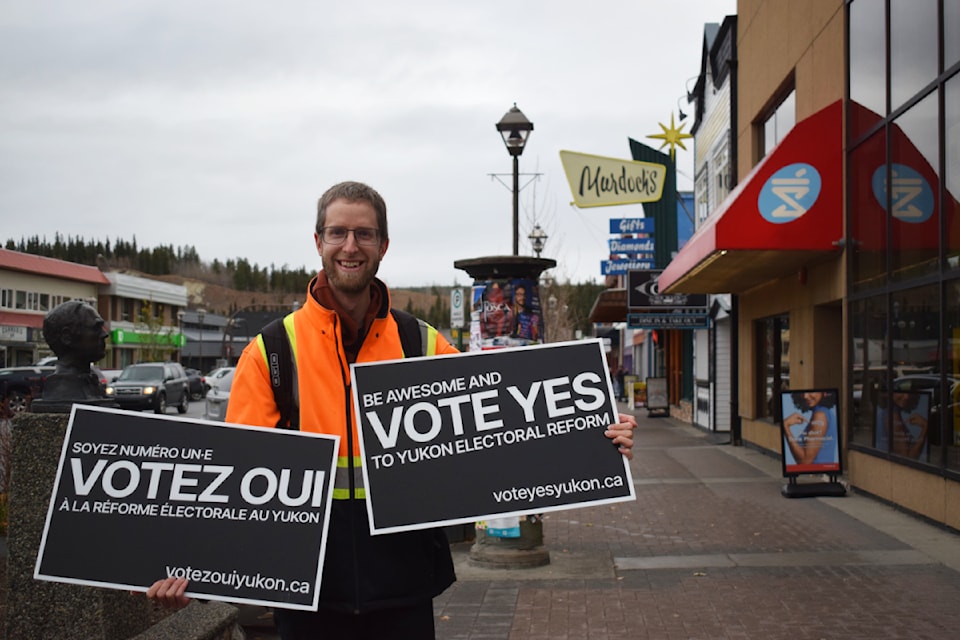 Sean standing in downtown Whitehorse holding two lawn signs, a “Votez OUI Yukon” sign and a “Vote YES Yukon” sign. Taken by Dana Hatherly from Yukon News.